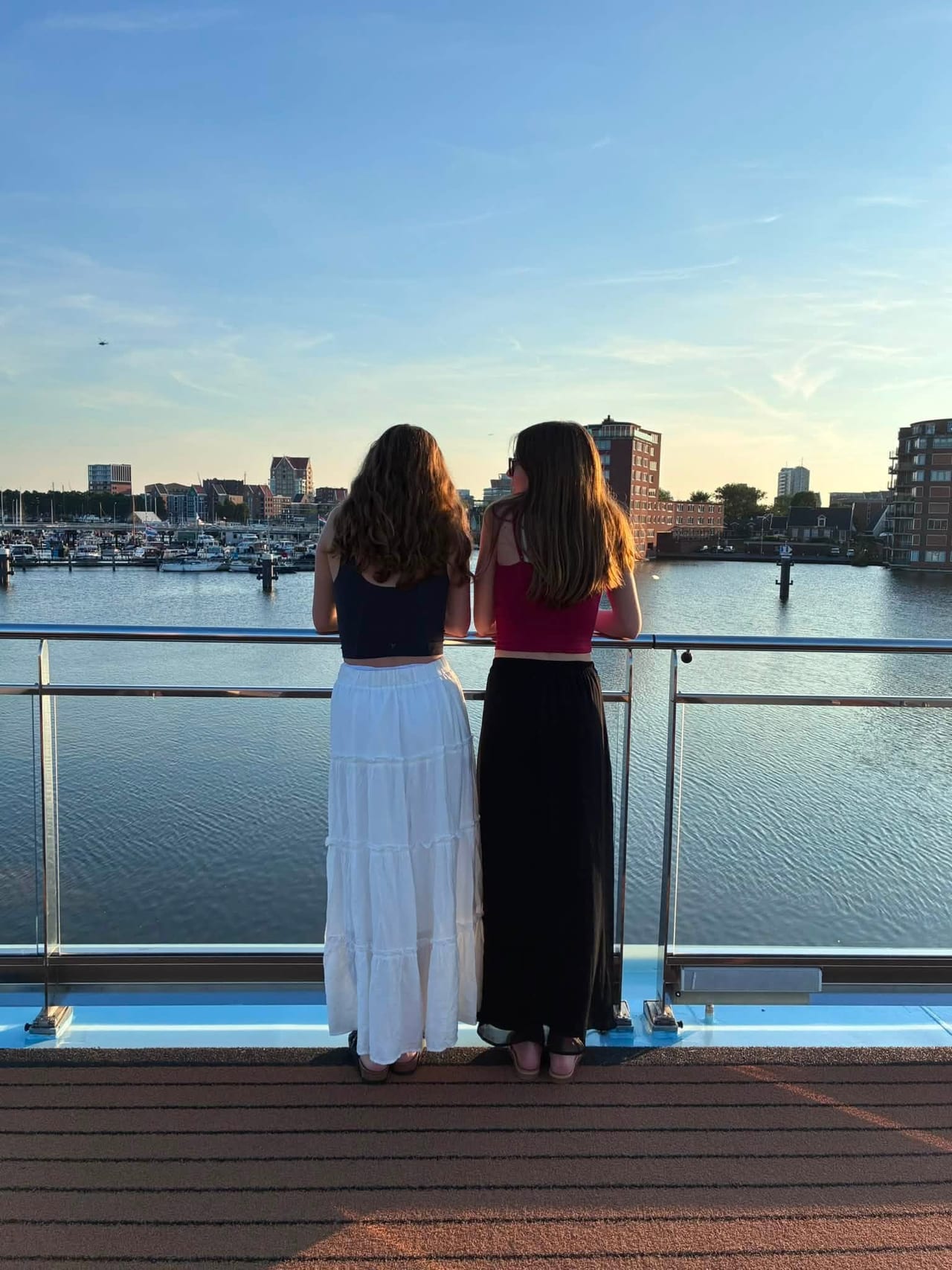 Bella and Abbi at the deck railing, backs to the camera, looking out over the harbor at sunset